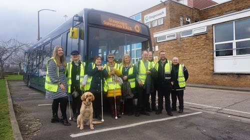 Gillmoss depot staff and RNIB volunteers in front of a Stagecoach Group bus displaying “Stagecoach Supporting R.N.I.B.” Several wear high-visibility vests and hold white canes, while others wear Stagecoach uniforms or lanyards. A guide dog sits at the front. The photo is taken in a parking area beside a brick building with a Stagecoach sign.  