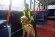 Image shows RNIB’s Area Campaigns Officer Terri Ballon wearing a high‑visibility yellow vest and a dark cap seated on a patterned blue‑and‑orange bus seat. In front of her sits a golden coloured Labrador Retriever guide dog wearing a white working harness looking directly at the camera. The bus interior features bright orange handrails, blue seats with an abstract orange pattern, and a window with railing faintly visible in the bright day outside.